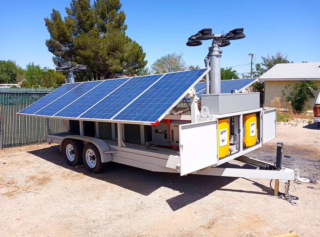 Dual-axle mobile power trailer with angled solar panels, open inverter bay, and LED light mast on a dirt lot
